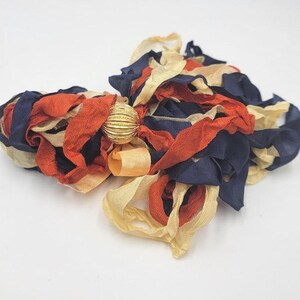 May include: A handmade hair accessory featuring fabric ribbons in navy blue, burnt orange, and cream colors. The ribbons are gathered and secured with a gold-colored, textured ball. This unique hair piece adds a touch of elegance.