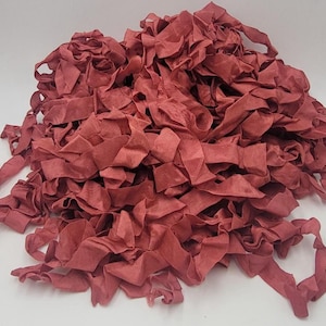 May include: A pile of ruffled, ribbon-like fabric in a muted, dusty rose color. The fabric appears to be soft and has a slightly shiny texture. The ribbons are layered and bunched together, creating a textured, voluminous appearance. This material could be used for crafting or decorative purposes.