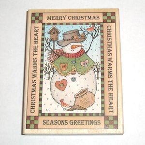 May include: A rectangular wooden rubber stamp featuring a snowman illustration with a hat, scarf, and birdhouse. The text reads MERRY CHRISTMAS, CHRISTMAS WARMS THE HEART, and SEASONS GREETINGS. The design includes a bird, a sack of bird seed, and hearts.
