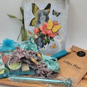 May include: A collection of craft supplies on a wooden tray. Includes a journal with a butterfly and floral design, fabric bundles, a blue pen, and small gift bags. The journal features a woman with butterfly wings.