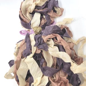 May include: A decorative tassel composed of layered fabric strips in cream, mauve, and brown hues. A gold-colored heart-shaped embellishment is centered on the tassel. The fabric strips are loosely arranged, creating a textured, layered effect.