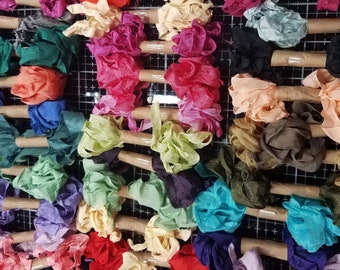 May include: Assortment of colorful fabric ribbon rolls, each tied with a brown paper band. The ribbons are arranged in rows on a black and white grid surface.