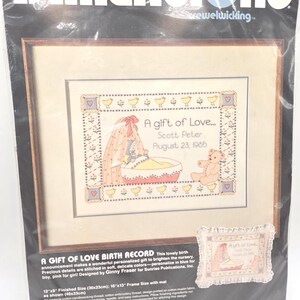 May include: A Dimensions crewelwicking birth record kit titled A Gift of Love featuring a baby in a cradle and a teddy bear. The design includes the text Scott Peter and August 23, 1985. The package indicates a finished size of 12 inches by 9 inches.