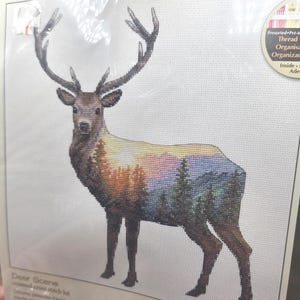 May include: A counted cross stitch kit titled Deer Scene featuring a stag with a double exposure design. The body of the deer displays a colorful landscape of orange and yellow trees, purple mountains, and green pine trees on white fabric.