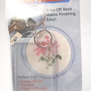 May include: A clear acrylic keychain kit in its original packaging. The packaging features the text Key Chain, Snap-Off Back Makes Finishing Easy!, and Perfect For Cross Stitch, Photos, Paper Artwork. A small cross stitch flower is visible.