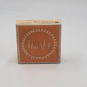 May include: A wooden stamp with the word "thanks" in white script, encircled by a white, swirly border, set against an orange background. The stamp is a cube shape, and the wood has a natural finish.