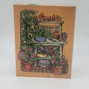 May include: A wooden block with a colorful illustration of a potting shed. The shed has a shelf with potted plants, a work table with tools, and a variety of flowers in pots.