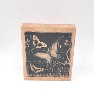 May include: A wooden stamp with a black rectangular design featuring three butterflies and decorative swirls. The butterflies are in various stages of flight, with detailed wing patterns. The stamp has a natural wood finish.