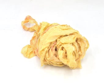 May include: A coiled length of pale yellow, semi-sheer fabric ribbon with a crinkled, textured surface. The ribbon features subtle, faint pinkish-orange color variations along its edges and folds, creating a soft, layered, and organic appearance.