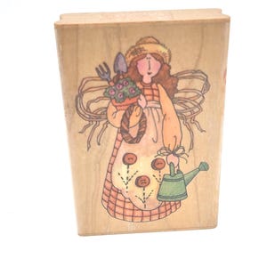 May include: A rectangular wooden rubber stamp featuring a line art illustration of a figure wearing a straw hat and apron, holding a potted plant with a trowel and fork, and carrying a green watering can. The design includes three buttons.