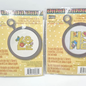 May include: Two cross-stitch kits featuring the words "LOVE" and "HOME" in yellow, with floral and heart accents. Each kit includes a 3.25-inch hoop, Aida fabric, floss, chart, and needle. The kits are packaged on a yellow background with white polka dots.