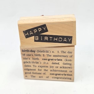 May include: Two wooden stamps. The top stamp reads "HAPPY BIRTHDAY" in black letters. The bottom stamp has a definition of "birthday" printed in black ink. The stamps are made of light-colored wood.