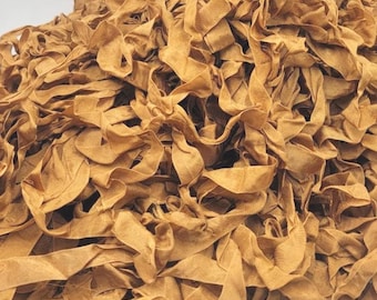 May include: A pile of tan-colored fabric strips, possibly ribbon or trim, with a slightly textured appearance. The strips are loosely arranged, creating a layered effect. The color is a warm, earthy tone.