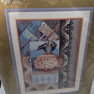 May include: Needlepoint kit featuring an intricate Native American design. The artwork includes a black and brown pot with a detailed pattern, set against a backdrop of geometric shapes and patterns in blue, brown, and white. The kit is packaged in a gold-colored plastic.