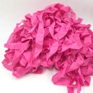 May include: A large, tangled pile of vibrant hot pink fabric ribbon strips. The material appears smooth and matte, with numerous individual strands folded and layered together in a dense, unstructured heap against a plain white background.