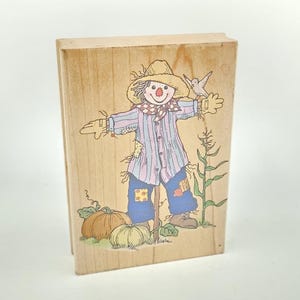 May include: A rectangular wooden rubber stamp featuring a line art illustration of a scarecrow wearing a straw hat, a striped shirt, and blue pants with patches. The design includes two pumpkins, a small bird, and a corn stalk in soft colors.