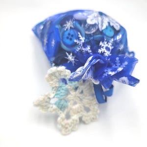May include: A blue organza drawstring pouch decorated with white snowflake patterns, filled with an assortment of blue buttons and beads. Attached to the drawstring is a small, six-pointed crocheted snowflake ornament in cream and light blue.