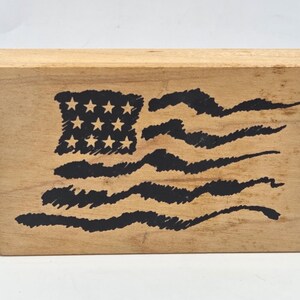 May include: A rectangular wooden rubber stamp featuring a black ink design of a stylized American flag. The flag graphic includes a square canton with twelve stars and four horizontal wavy stripes, all rendered in a hand-drawn, textured black ink style.