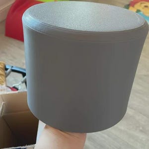 May include: A gray, cylindrical container with a textured top and a slightly tapered body. The container is held by a hand, and the background includes a cardboard box and a red object.