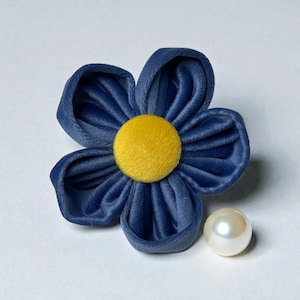 May include: A blue velvet flower with a yellow center and a pearl. The flower has five petals and a round, yellow center. The pearl is white and round. The flower and pearl are on a white background.