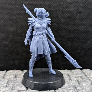 Może przedstawiać: Szara, trójwymiarowa figurka wojowniczki trzymająca włócznię. Figurka jest ubrana w zbroję i spódnicę, ma też kucyk. Figurka stoi na okrągłej podstawie.