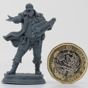 May include: A grey miniature figurine of a musician playing a hurdy-gurdy, wearing a long coat and hat. A one-pound coin is next to the figurine. The figurine is approximately 5 cm tall.