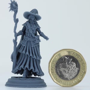 Op de afbeelding: Grijze miniatuurfiguur van een fantasiekarakter, mogelijk een tovenaar of tovenares, die een staf vasthoudt. De figuur staat naast een Britse munt van één pond voor de schaal. De figuur draagt een hoed en kleding in lagen.