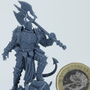 Puede incluir: Figura en miniatura gris detallada de un guerrero con armadura adornada, sosteniendo una gran hacha de doble hoja. La figura tiene cuernos y una cola. Una moneda de una libra está al lado de la figura para la escala.