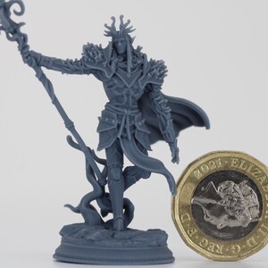 Puede incluir: Figura en miniatura gris detallada de un personaje de fantasía, sosteniendo un bastón y vistiendo una armadura y una capa. La figura está junto a una moneda de una libra británica para la escala. La figura mide unos 5 cm de altura.