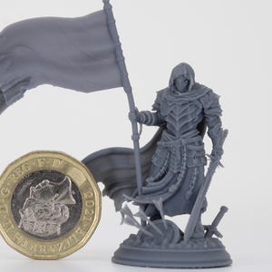 Puede incluir: Figura en miniatura gris de un guerrero encapuchado que sostiene una bandera y dos espadas, de pie sobre una base con espadas y flechas rotas. Una moneda de una libra está al lado de la figura para la escala.