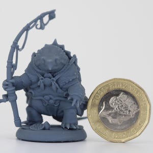 Könnte beinhalten: Detaillierte, graue Figur einer froschartigen Kreatur, die eine Angelrute hält. Die Figur befindet sich neben einer britischen Ein-Pfund-Münze, die als Größenreferenz dient. Die Kreatur trägt ein detailliertes Outfit.