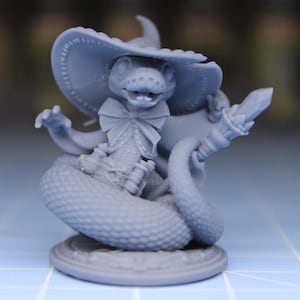 Puede incluir: Figura gris de una serpiente con sombrero de bruja y pajarita, sosteniendo un bastón con un cristal. La serpiente está enrollada sobre una base redonda. La figura tiene escamas detalladas y es de estilo caprichoso.