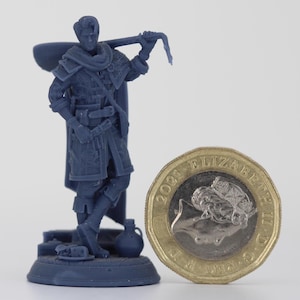 May include: A blue miniature figurine of a character, possibly for a tabletop game, with a tool over their shoulder. A one-pound coin is next to the figurine for scale.