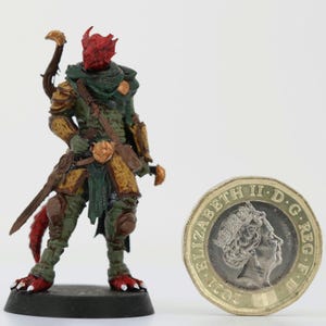 Può includere: Miniatura di una creatura umanoide fantasy con pelle rossa e corna, vestita con un'armatura verde e oro, che tiene una spada. Accanto c'è una moneta da una sterlina britannica per la scala.