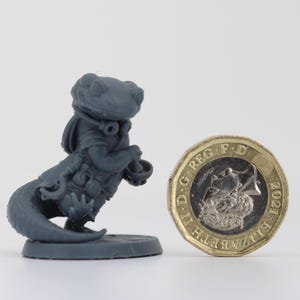 以下が含まれることがあります： 小さなボウルを持った、トカゲのような生き物、おそらくファンタジーキャラクターの灰色のミニチュアフィギュア。フィギュアは、スケールを示すために英国の1ポンド硬貨の隣にあります。フィギュアは円形のベースにあります。