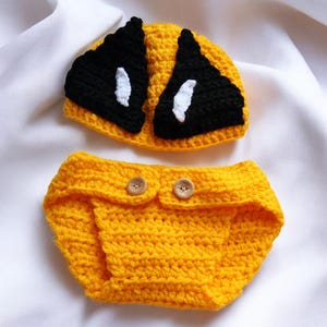 May include: A yellow crocheted baby outfit with a matching hat. The hat has black and white eyes.