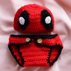 May include: Red and black crocheted baby hat and diaper cover with white eye details. The hat has a black eye mask with a white circle in the center. The diaper cover has two brown buttons.