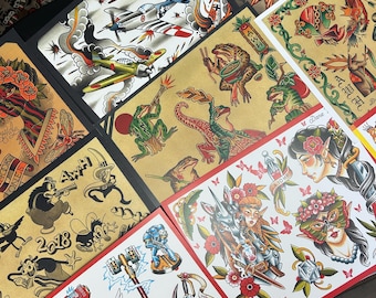 May include: A collection of tattoo flash art prints featuring various traditional tattoo designs. The prints showcase colorful illustrations of airplanes, frogs, flowers, and mythical creatures. Some prints include text such as "SHOOK ONES" and "Magic Mirror".