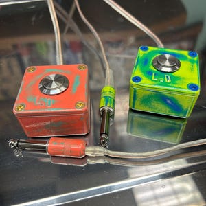 May include: Two square, box-shaped foot switches with metallic push buttons on top. One box is orange with teal accents, and the other is lime green with blue swirls. Each features the letters L.D. and is connected by clear cables to 1/4-inch jacks.