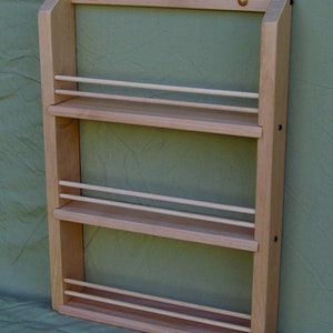 May include: A wooden spice rack with three shelves and a light wood finish. The rack is mounted on a wall and has a simple design.