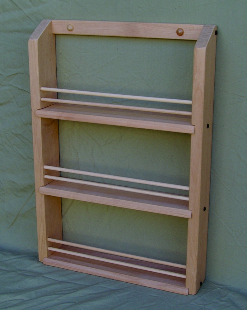 May include: A wooden spice rack with three shelves and a light wood finish. The rack is mounted on a wall and has a simple design.