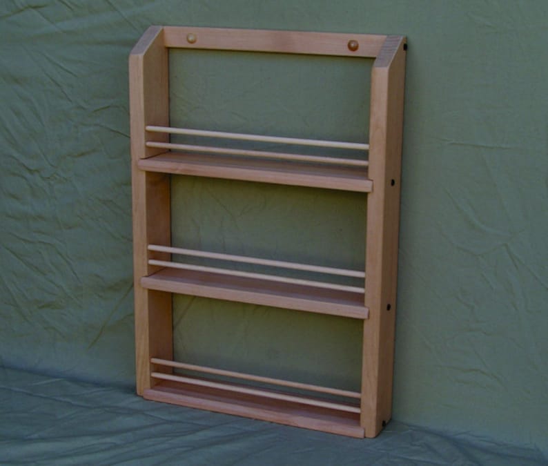 May include: A wooden spice rack with three shelves and a natural finish. The rack is mounted on a wall and has a simple design.