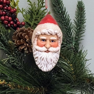 May include: A white ceramic Santa Claus ornament with a red hat and a white beard. The ornament is hanging from a red string.