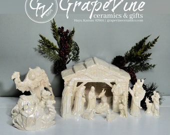 Puede incluir: Un belén de cerámica con acabado iridiscente. El conjunto incluye figuras de la Sagrada Familia, los Reyes Magos, animales y un establo. El logotipo de GrapeVine Ceramics está en la parte superior. La escena está rodeada de vegetación decorativa.