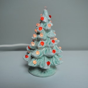 May include: A small, ceramic Christmas tree with a light blue glaze and red lights. The tree is lit up and has a white cord.