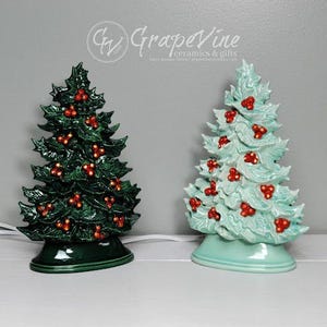 May include: Two ceramic Christmas tree figurines. One is green with red berries and the other is light blue with red berries. Both are on a white surface.