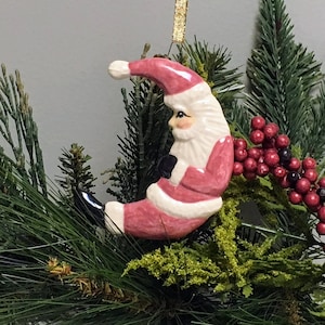 May include: A pink and white ceramic Christmas ornament shaped like a crescent moon with Santa Claus sitting on it. Santa is wearing a red and white suit with a black belt and a white beard. The ornament is hanging from a gold string.