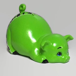 May include: A green ceramic piggy bank with black and white details. The piggy bank is lying on its side with its head resting on the ground. It has blue eyes and a small, curled tail.