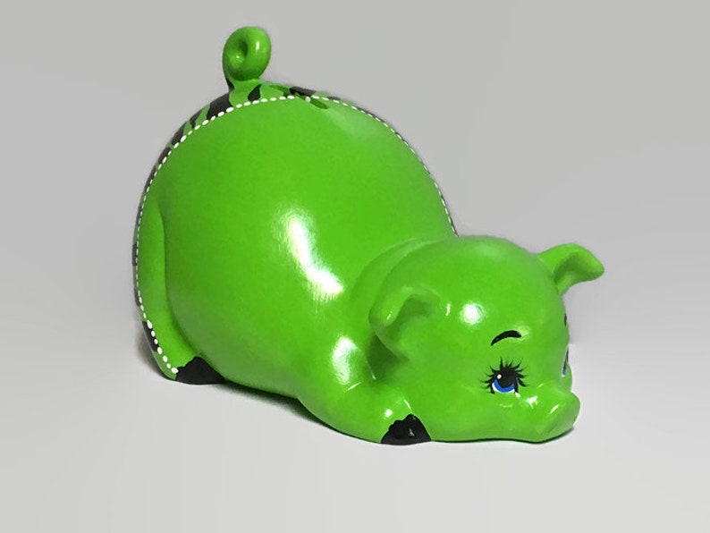May include: A green ceramic piggy bank with black and white details. The piggy bank is lying on its side with its head resting on the ground. It has blue eyes and a small, curled tail.
