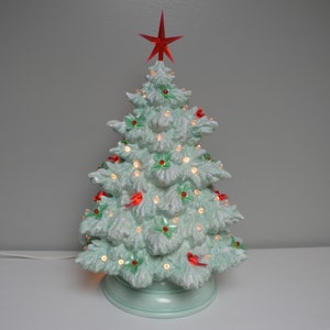 Op de afbeelding: Een kerstboom van keramiek met een lichtgroene glazuur, versierd met rode kardinaals, groene hulst en witte lampjes. De boom heeft een rode ster bovenop en een wit snoer.